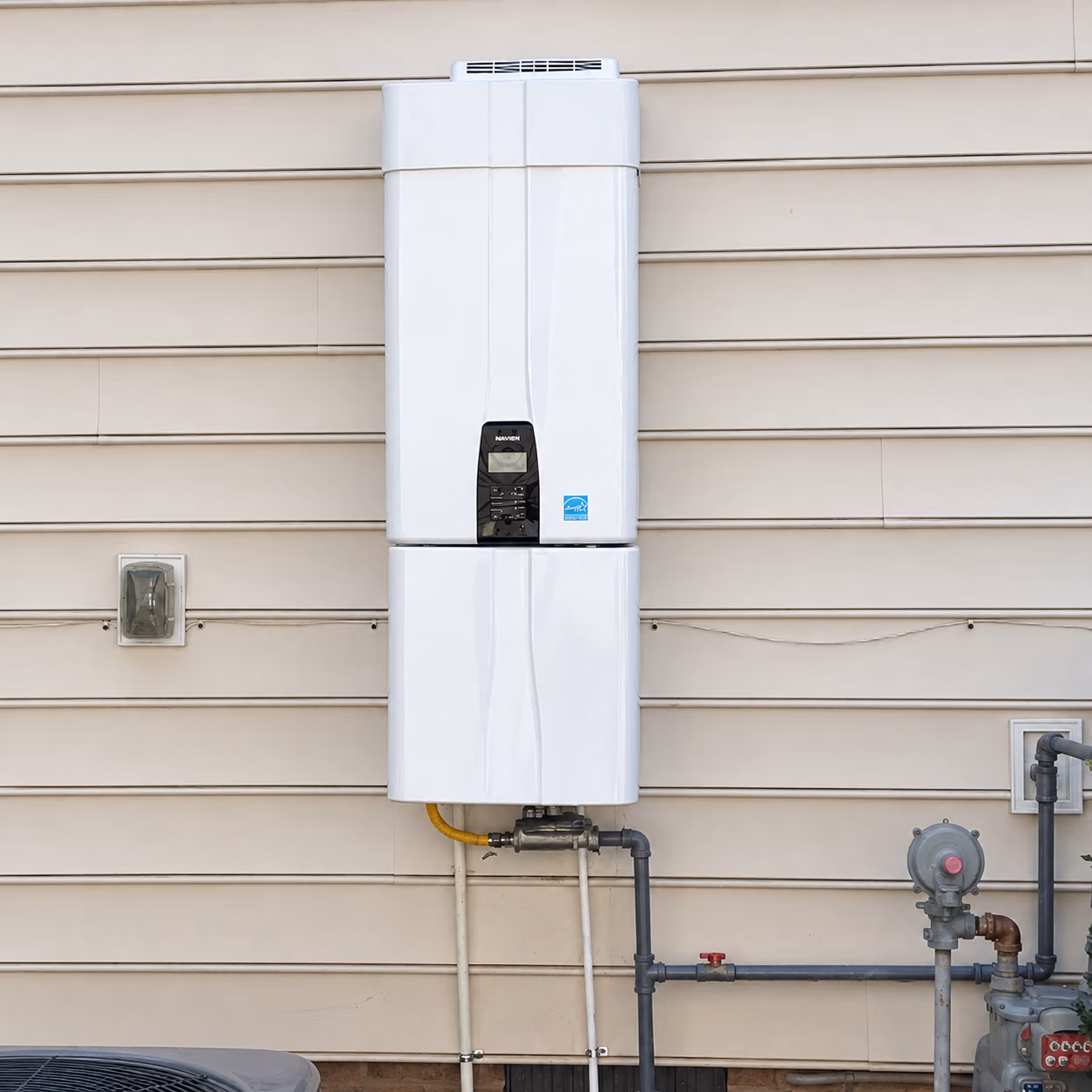 White tankless water heater unit mounted on beige siding with pipes and valves below.