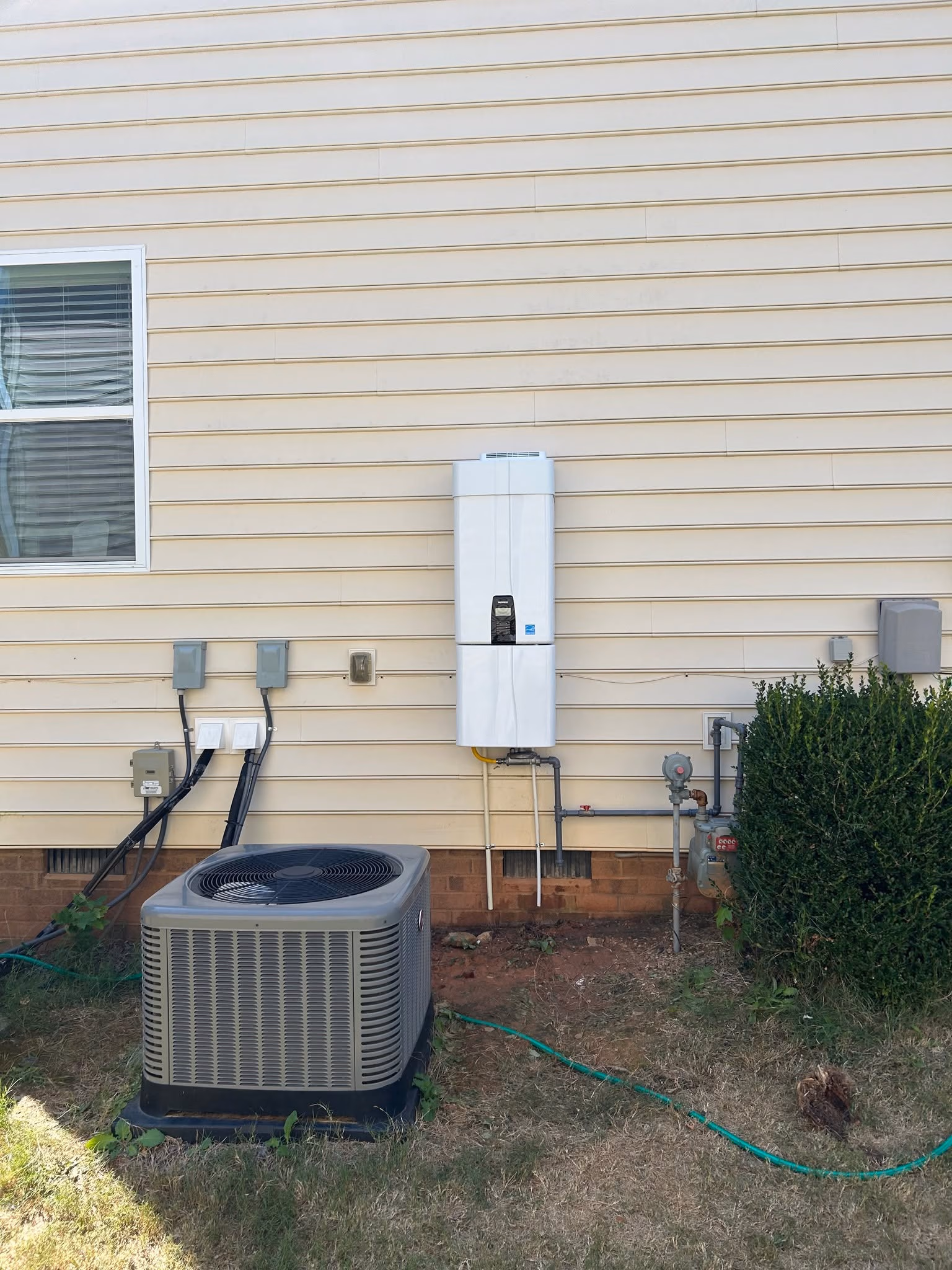 Outdoor HVAC unit and gas meter mounted on beige house siding with window and green bush nearby.