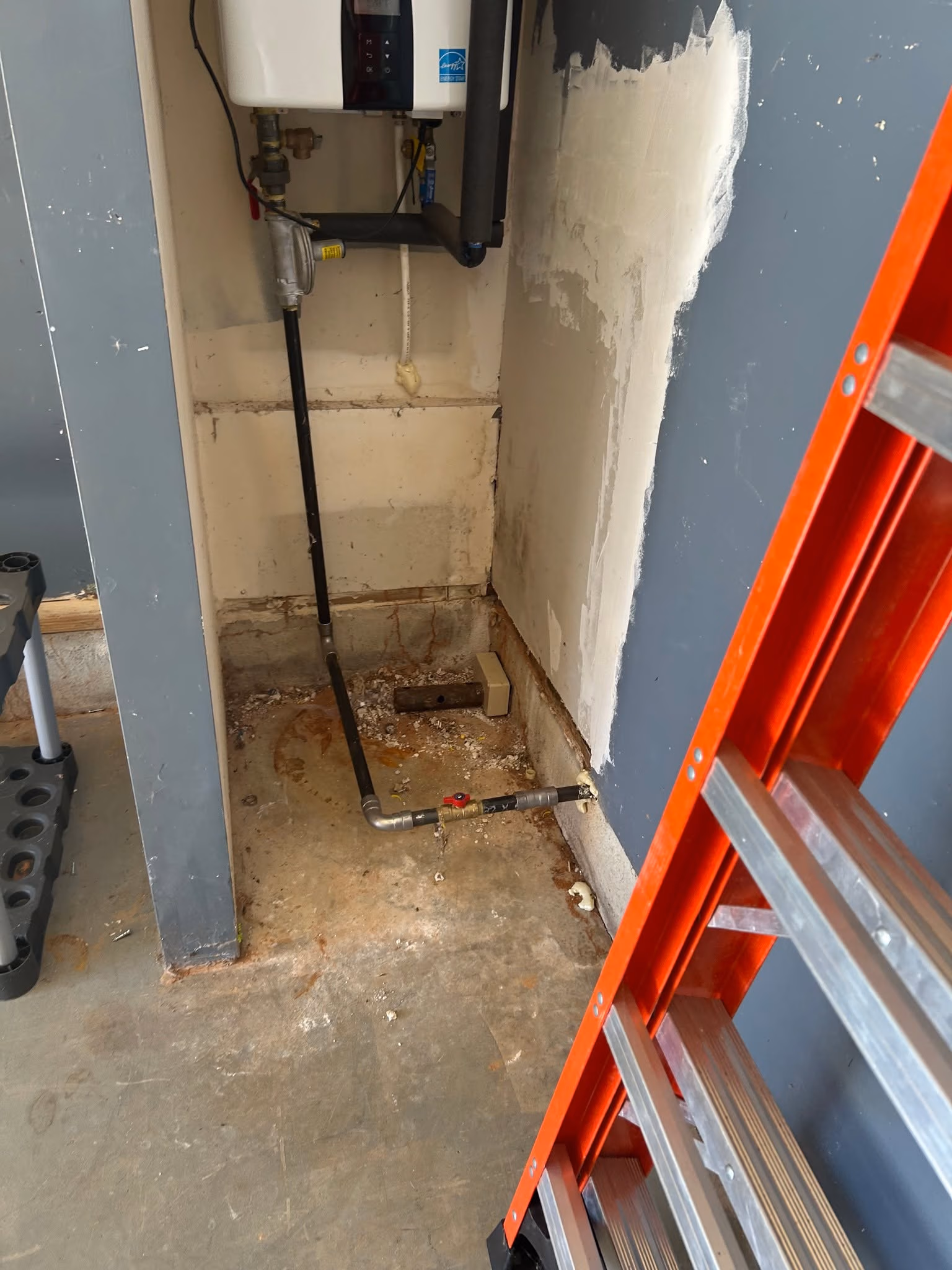 Corner of a room with exposed pipes, a wall unit, a red ladder, and a concrete floor with stains.