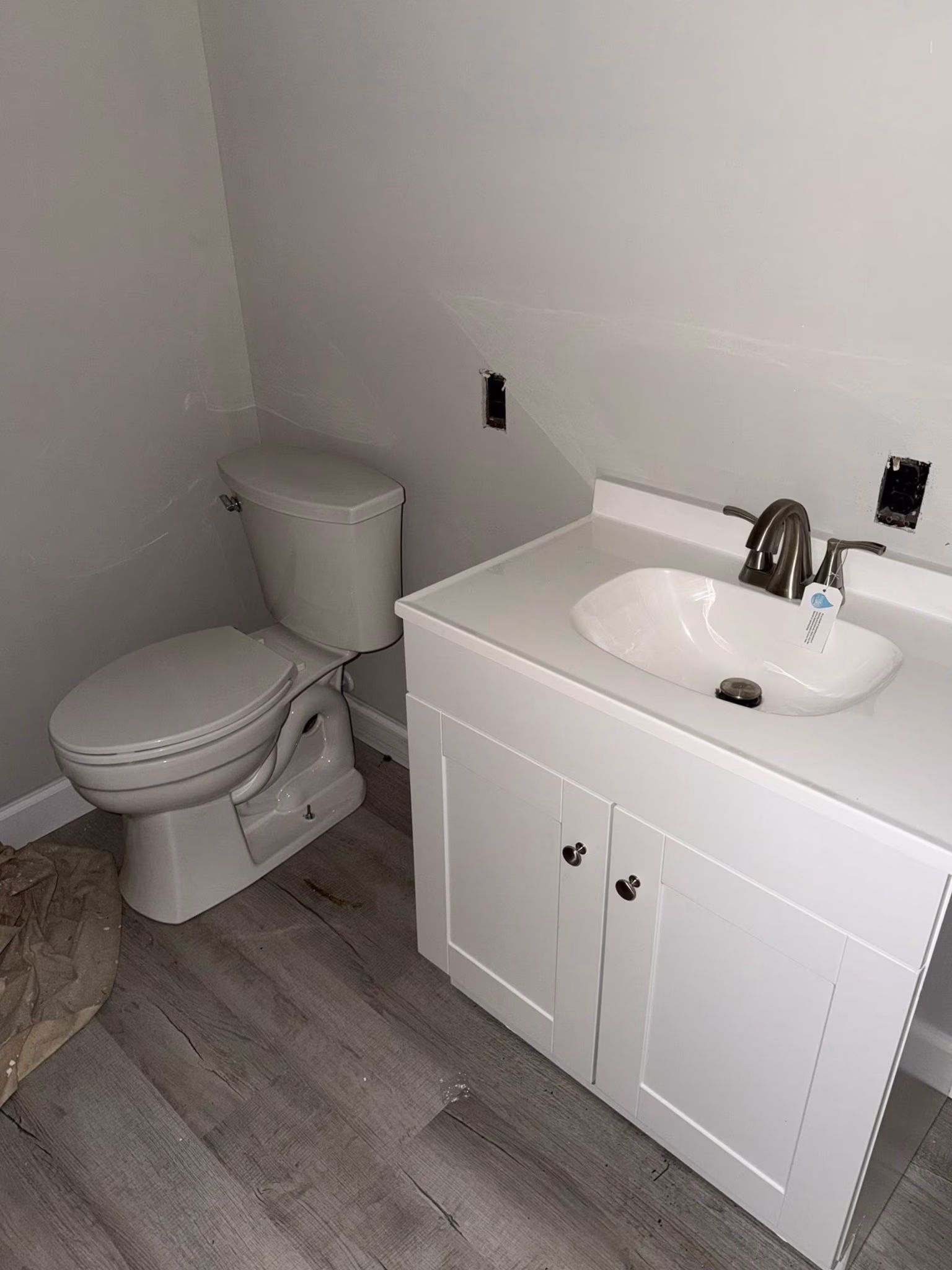 Small bathroom with white toilet, white vanity with sink, gray wood floor, and bare light switch boxes.