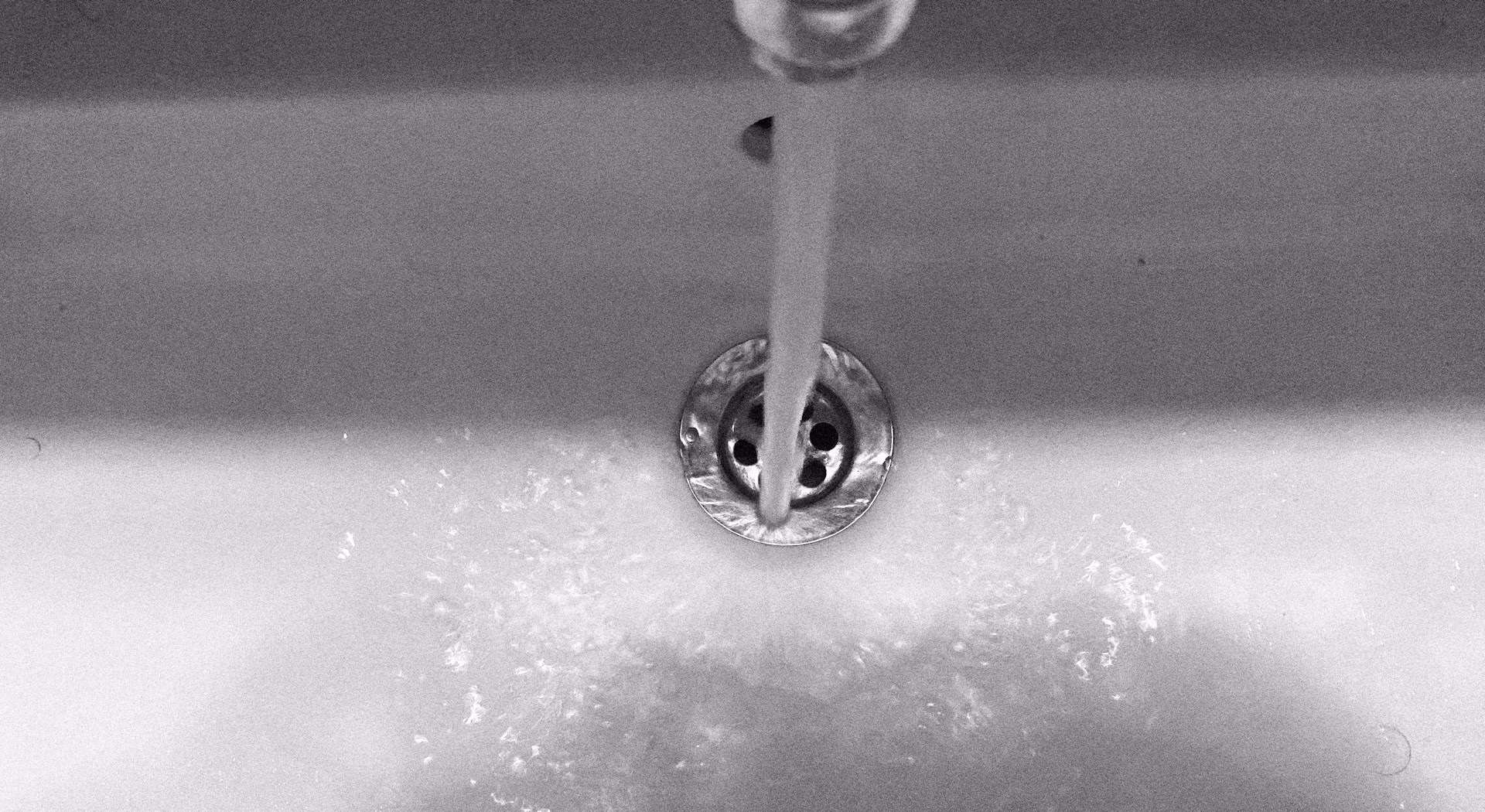 Stream of water flowing from faucet into a white sink drain with metal grate.