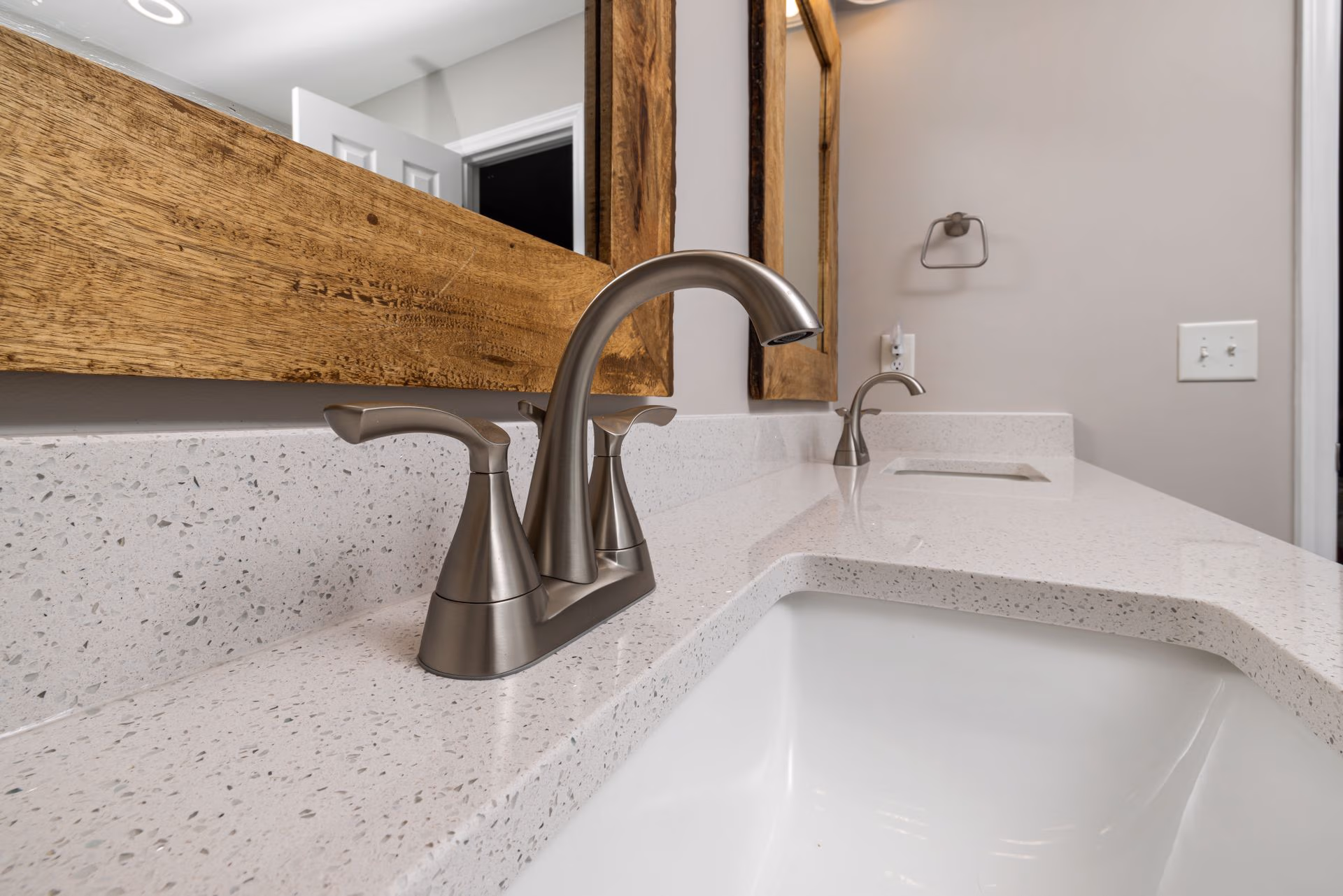 Close-up of a modern bathroom sink with silver faucet and wooden framed mirror on a white speckled countertop.