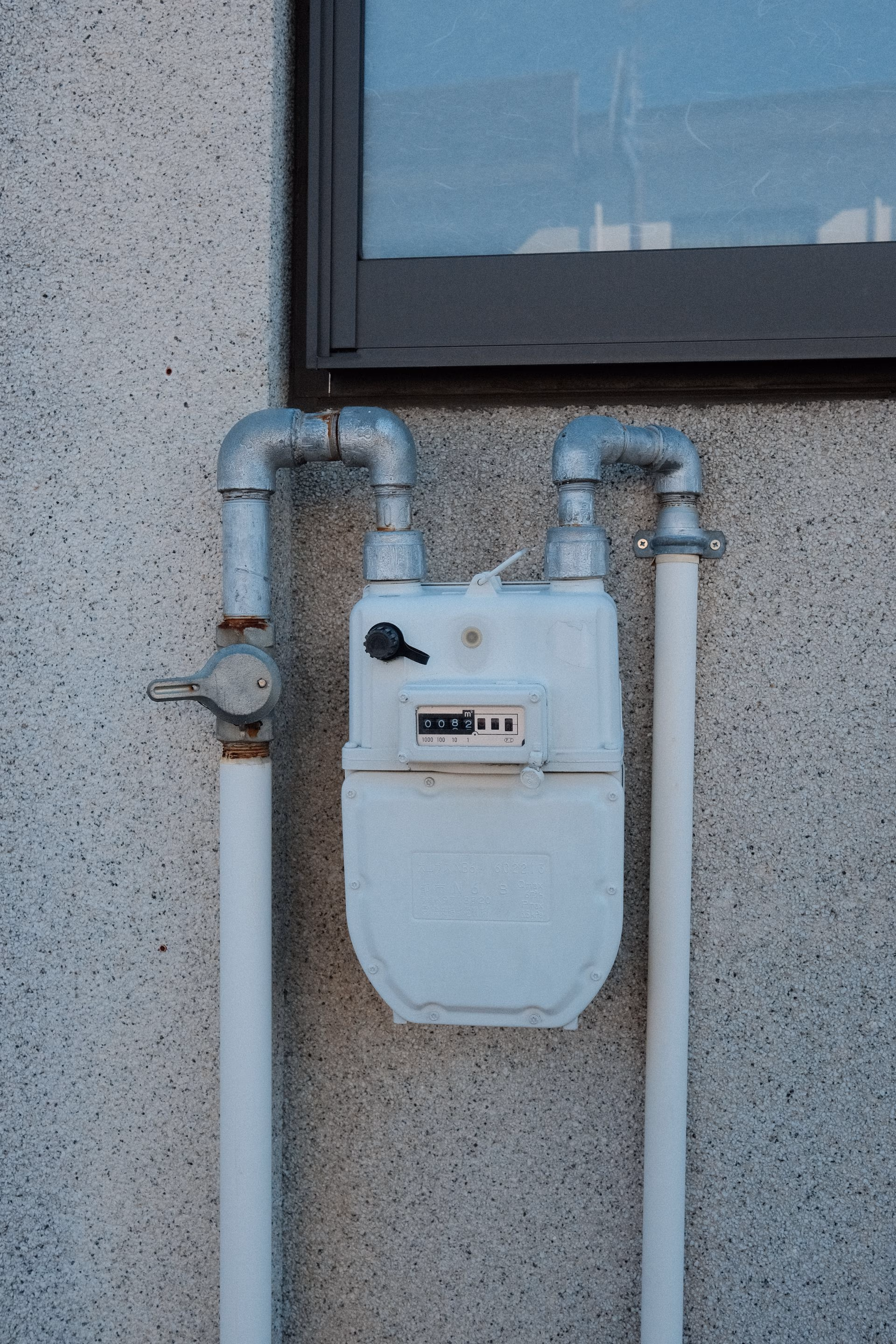White gas meter with pipes attached to a textured exterior wall beneath a window.