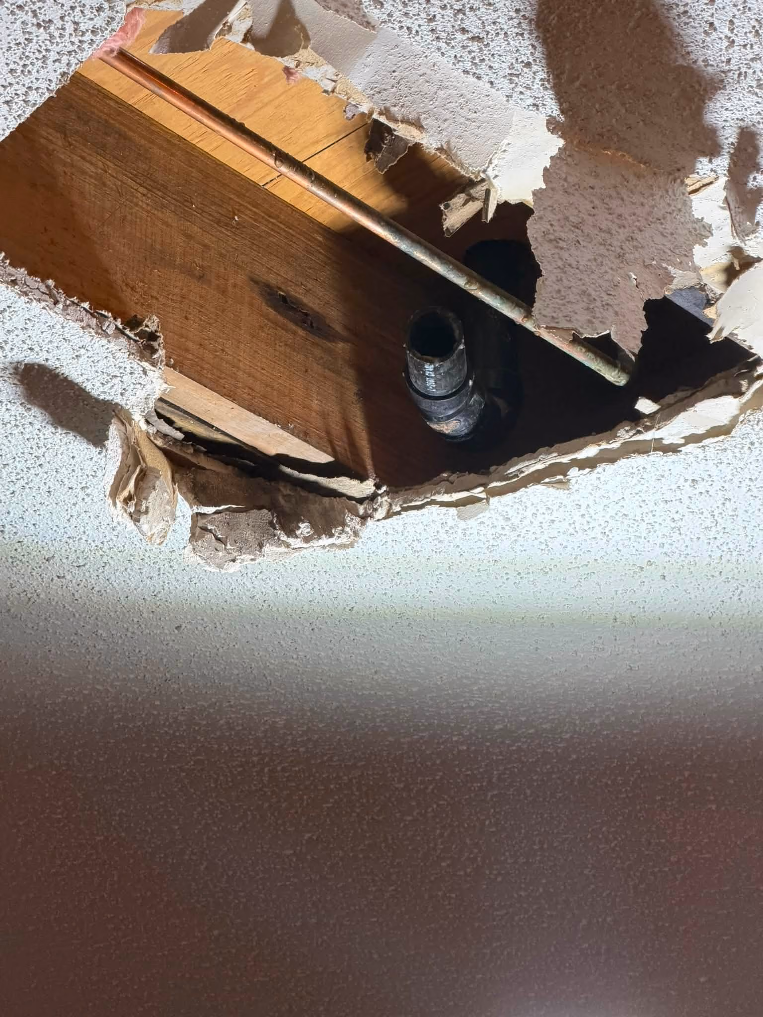 Ceiling hole exposing wooden beam, copper pipe, and black plastic plumbing pipe inside.