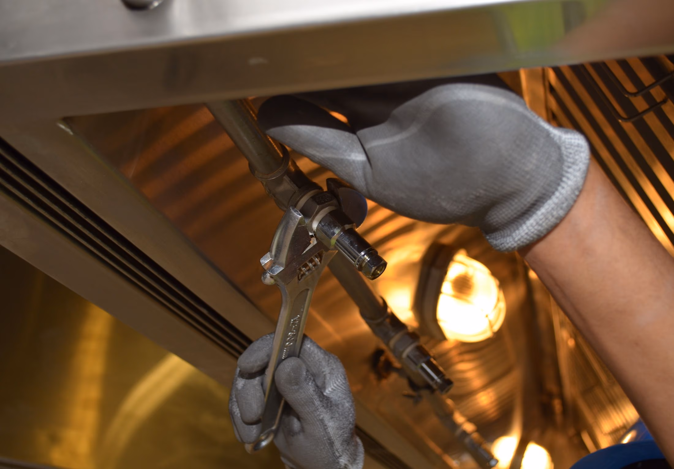 Gloved hands using a wrench to tighten a metal pipe fitting under a reflective surface.