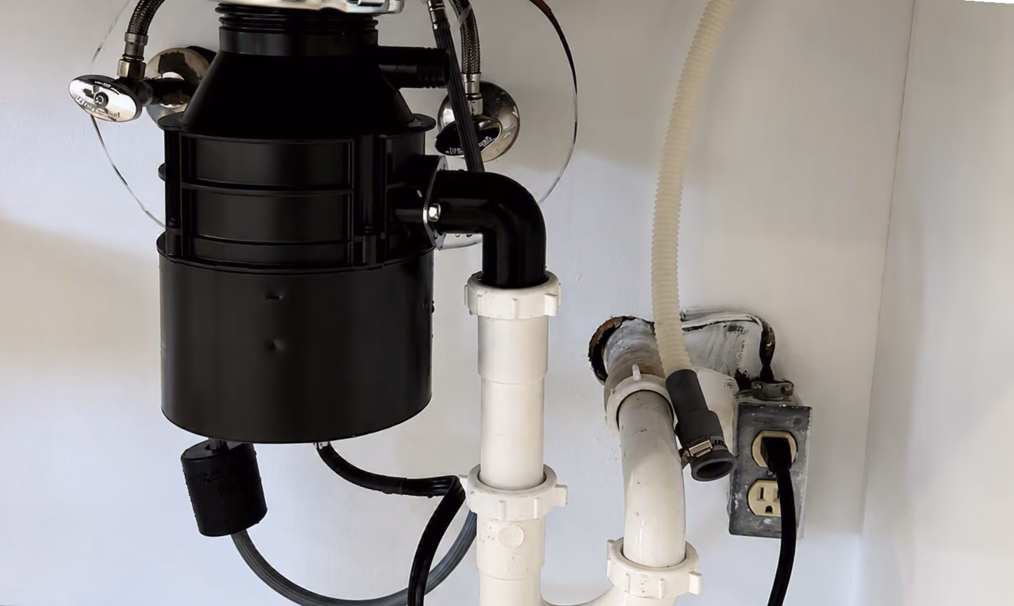 Under-sink plumbing with black garbage disposal unit, white pipes, and power outlet on wall.