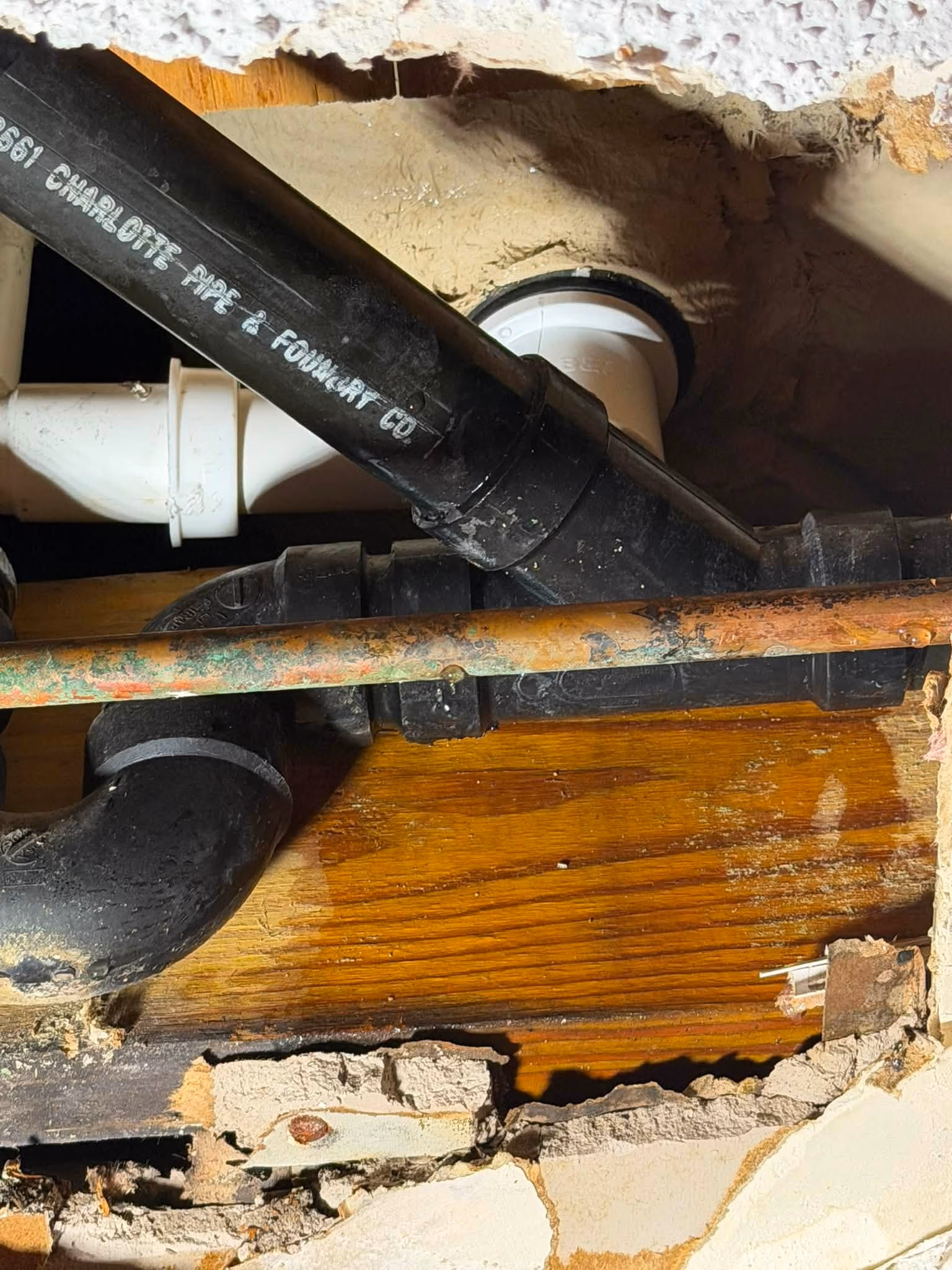 Close-up of black and white plumbing pipes and a corroded copper pipe behind a wall opening.