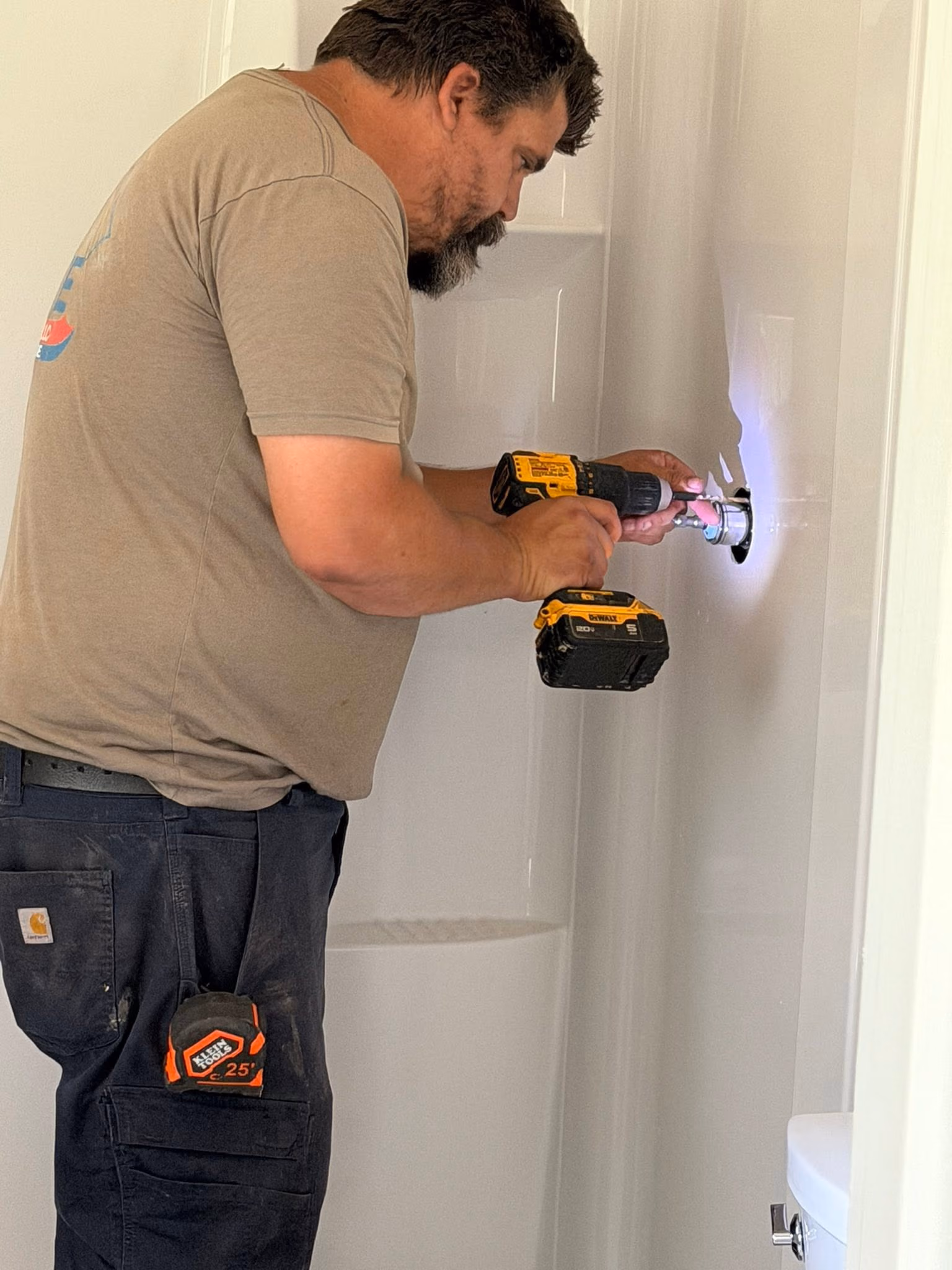 Man using cordless drill to install or repair plumbing in a white shower stall.