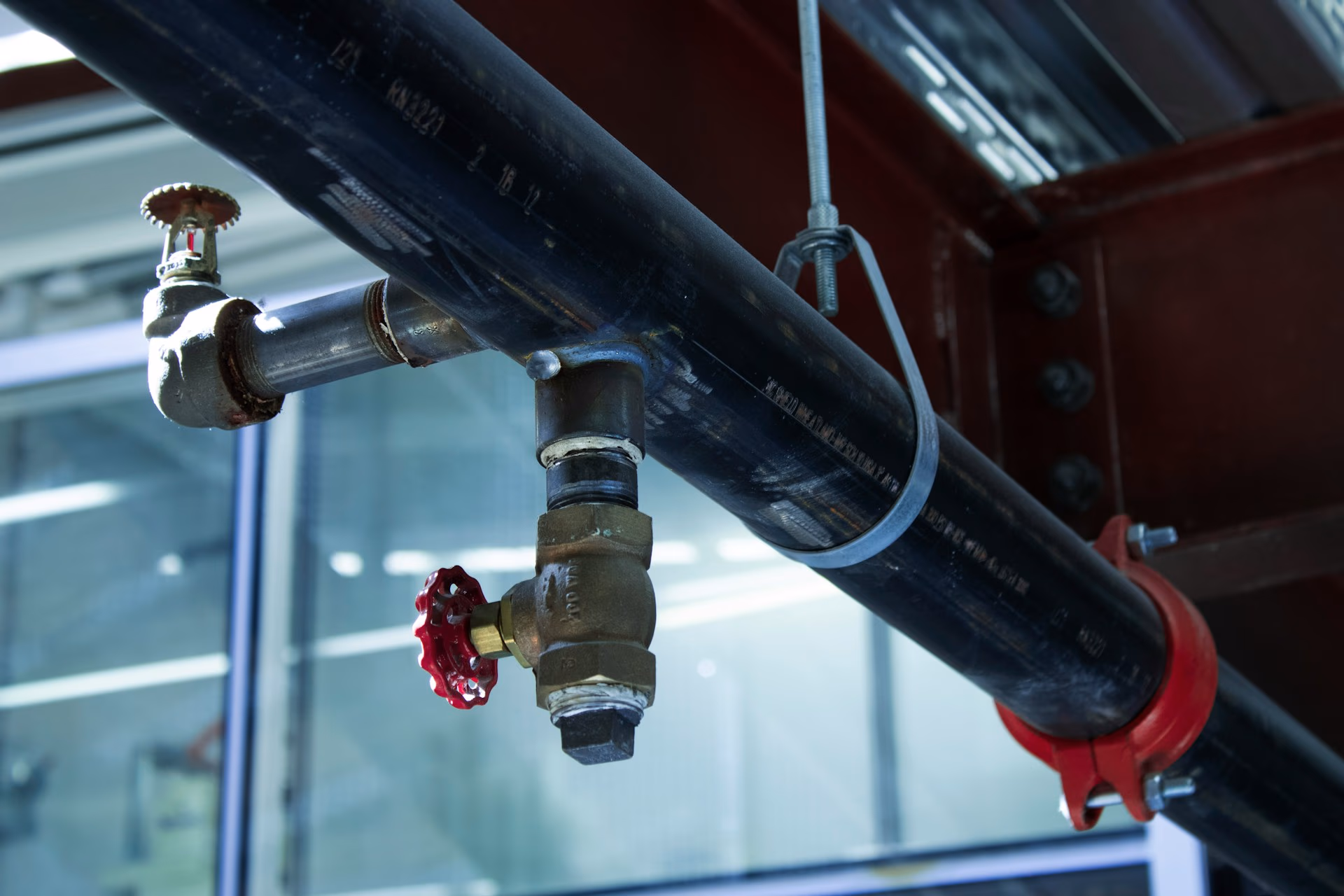 Close-up of industrial black pipe with valves and red clamps inside a building.