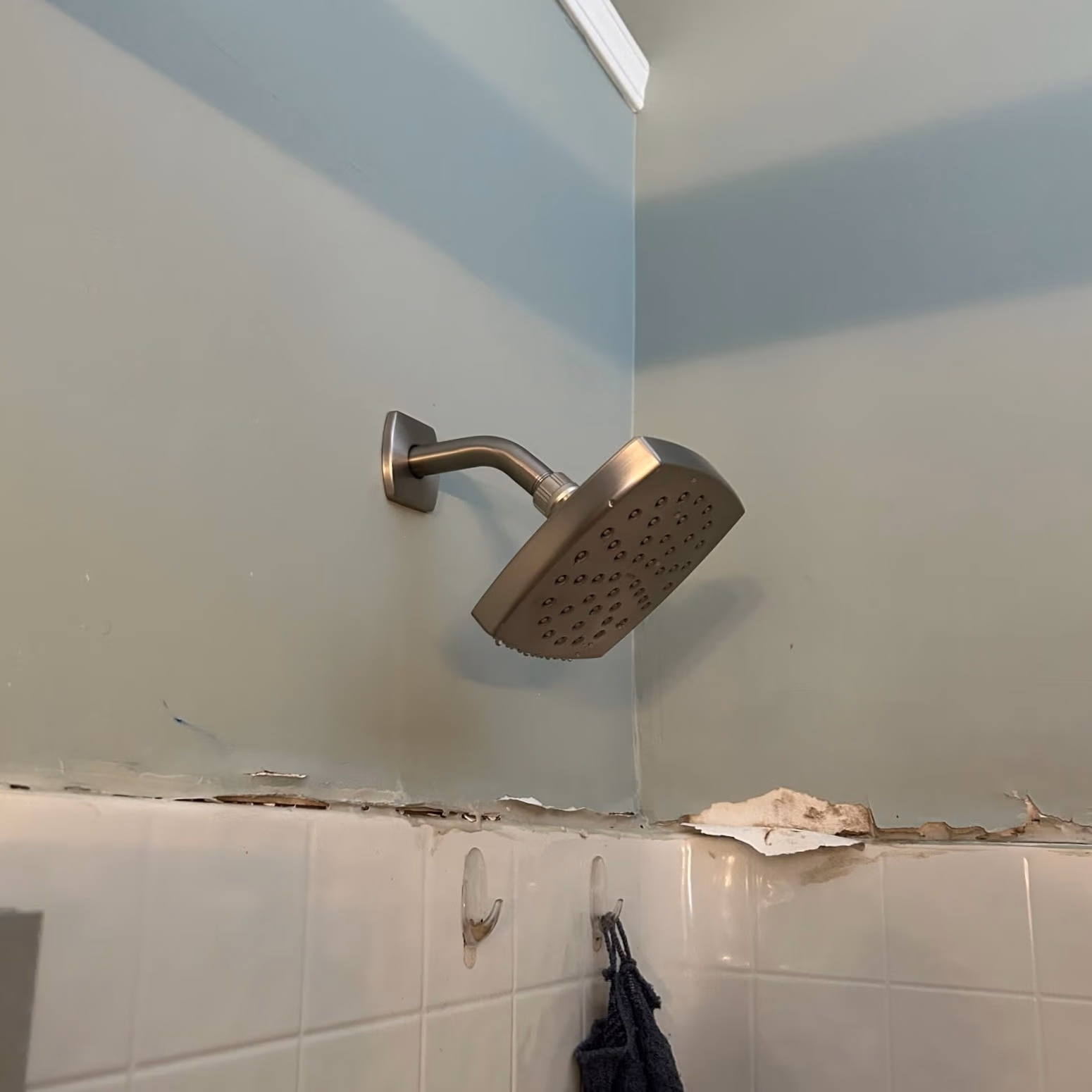 Metallic square shower head mounted on a light blue bathroom wall above damaged paint and white tiles.
