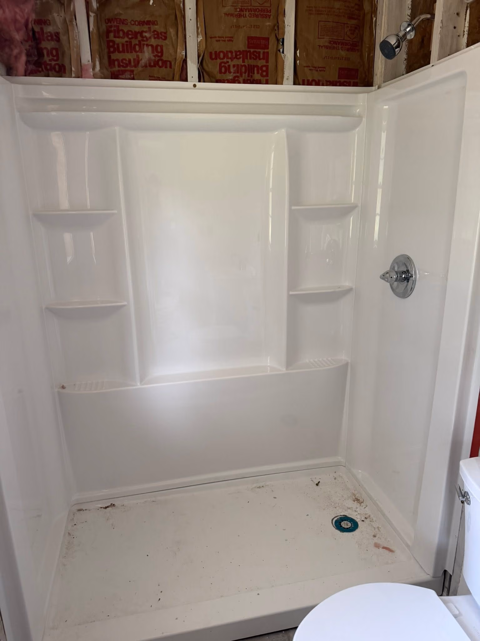 White shower stall with built-in shelves, chrome showerhead, and exposed insulation above.