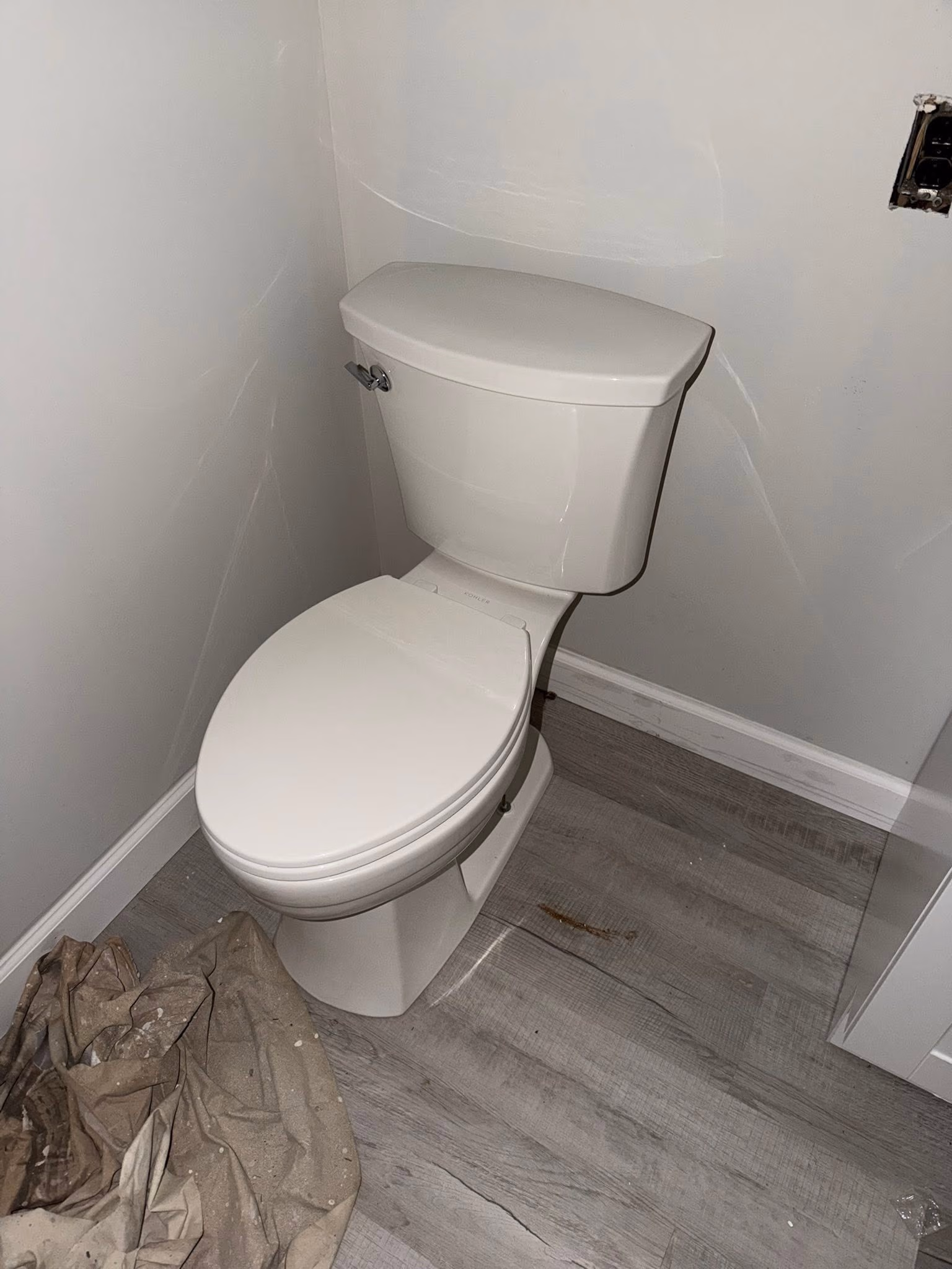 White closed toilet in a corner with gray wood floor and a cloth on the floor.